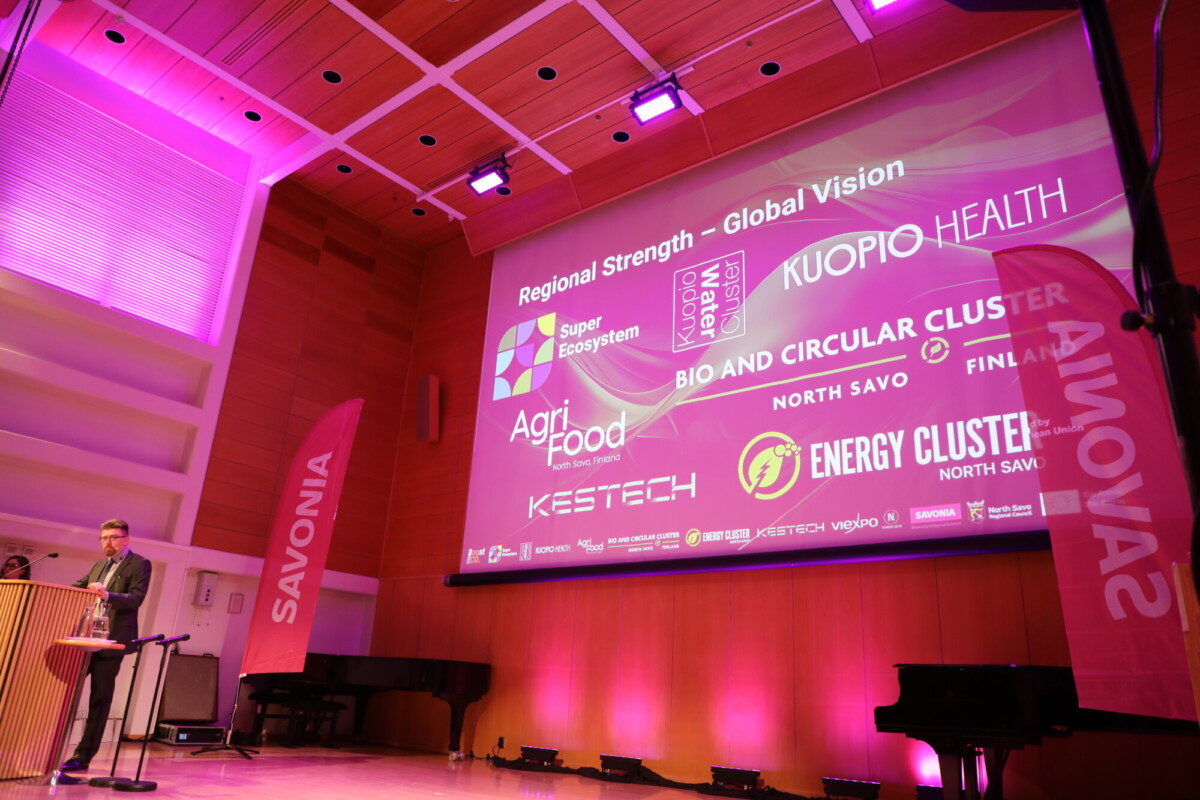 Henkilö seisoo korokkeella näyttämöllä, jossa on suuri näyttö, jolla näkyy vaaleanpunaisessa ja violetissa valaistuksessa logoja ja tekstiä alueellisista innovaatioklustereista. Savonia näkyy banderollissa ja höyhenlipussa.