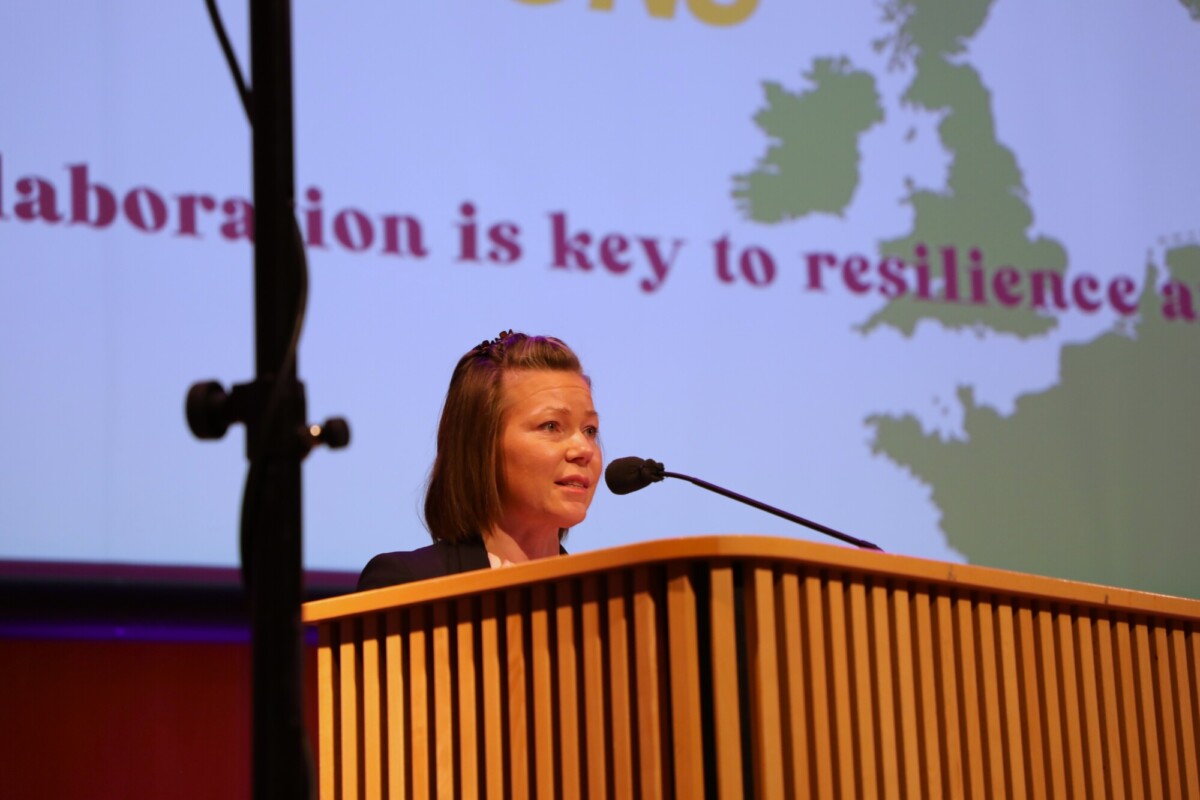 Nainen puhuu korokkeella, jossa on mikrofoni. Hänen takanaan näytöllä näkyy Ison-Britannian kartta ja osittainen teksti: Collaboration is key to resilience.