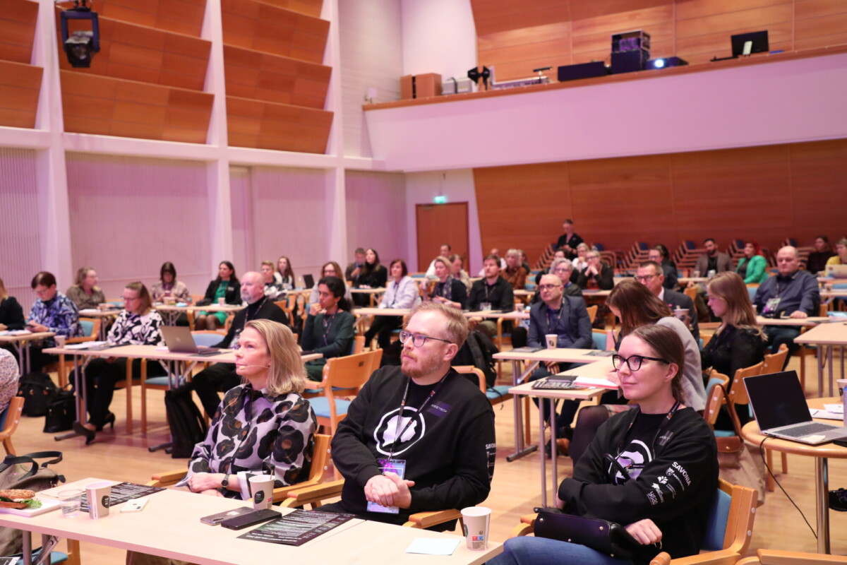 Joukko ihmisiä istuu pöydissä suuressa kokoushuoneessa ja kuuntelee tarkkaavaisesti esitystä. Joillakin on kannettavat tietokoneet ja muistikirjat. Ilmapiiri on ammattimainen ja sitoutunut.