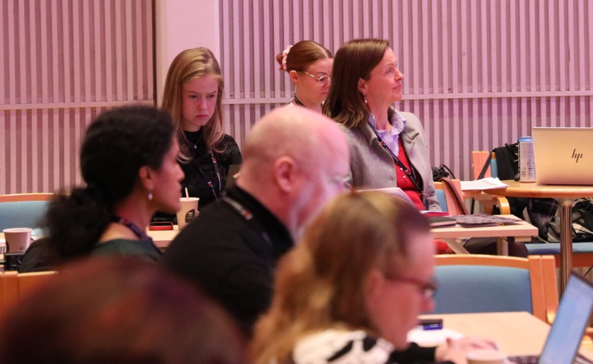 Joukko ihmisiä istuu neuvotteluhuoneen pöydissä ja kuuntelee tarkkaavaisesti. Jotkut käyttävät kannettavia tietokoneita, kun taas toiset keskittyvät puhujaan. Ilmapiiri vaikuttaa sitoutuneelta ja ammattimaiselta.