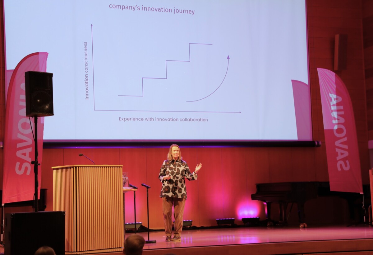 Nainen esittelee lavalla yrityksen innovaatiomatka -nimistä kuvaajaa, taustalla vaaleanpunaisia SAVONIA-banderolleja ja näyttämövalaistus.