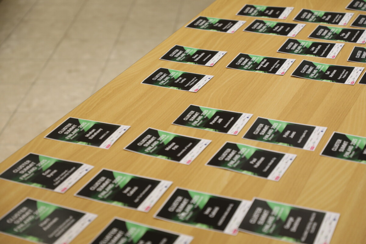 Puinen pöytä, jossa on useita mustavihreitä tapahtuman nimikylttejä, jotka on aseteltu riveihin, ja jokaisessa kyltissä on valkoinen teksti. Nimikyltit on järjestetty ja valmiina jakelua varten.