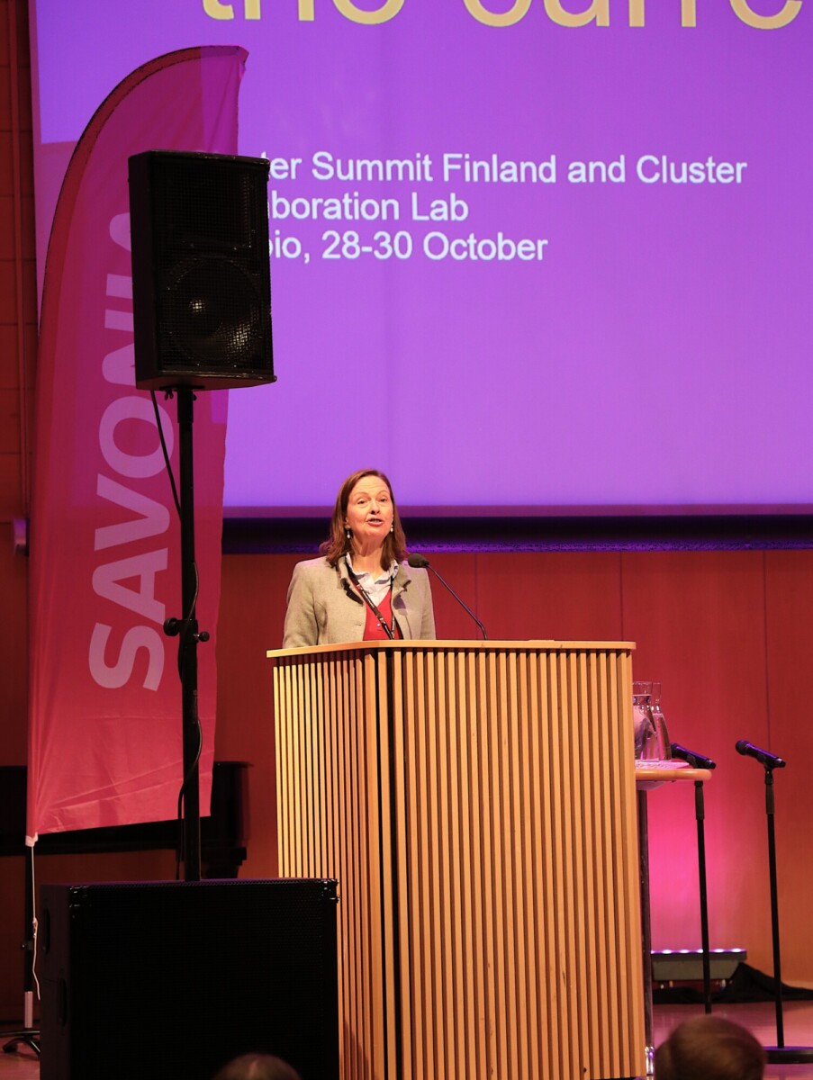 Nainen seisoo puisen korokkeen ääressä ja puhuu lavalla violetissa valaistuksessa. Hänen takanaan näytöllä näkyy tietoa Suomessa järjestettävästä huippukokouksesta. Myös vaaleanpunainen "Savonia"-banderolli ja äänentoistolaitteet ovat näkyvissä.