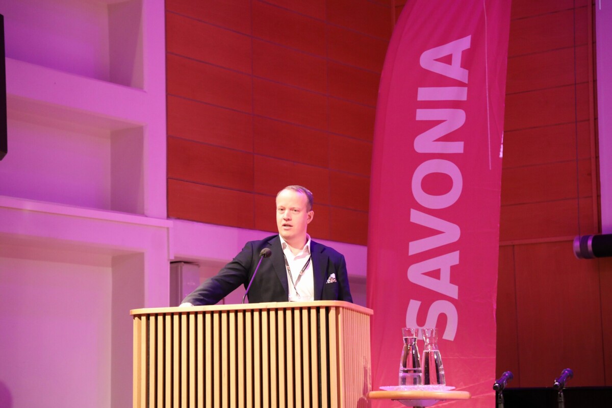 Pukumies seisoo puisella korokkeella ja puhuu lavalla. Hänen takanaan on korkea vaaleanpunainen banderolli, jossa lukee valkoisin kirjaimin SAVONIA. Korokkeen vieressä olevalla pienellä pöydällä on kaksi vesikannua ja lasi.