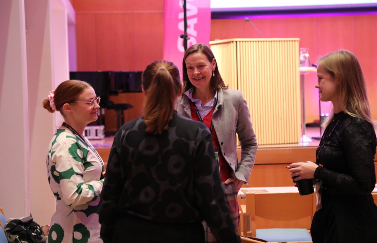 Neljä naista seisoo puoliympyrässä keskustelemassa kirkkaasti valaistussa tapahtumatilassa, jossa on puiset seinät ja taustalla koroke. Yhdellä naisella on muki kädessään, ja he näyttävät verkostoituvan tai seurustelevan.