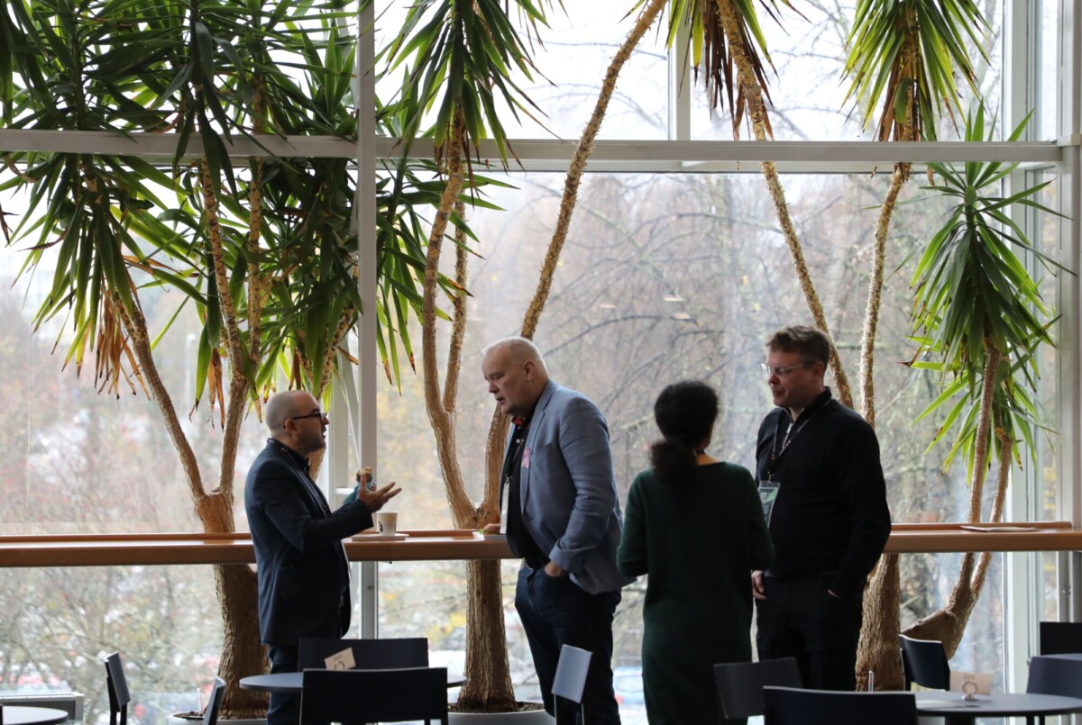 Neljä ihmistä seisoo ja keskustelee valoisassa sisätilassa, jossa on suuret ikkunat ja korkeat ruukkukasvit. Kaksi miestä keskustelee toisiaan vasten, kun taas mies ja nainen seisovat vieressä. Etualalla näkyy pöytiä ja tuoleja.