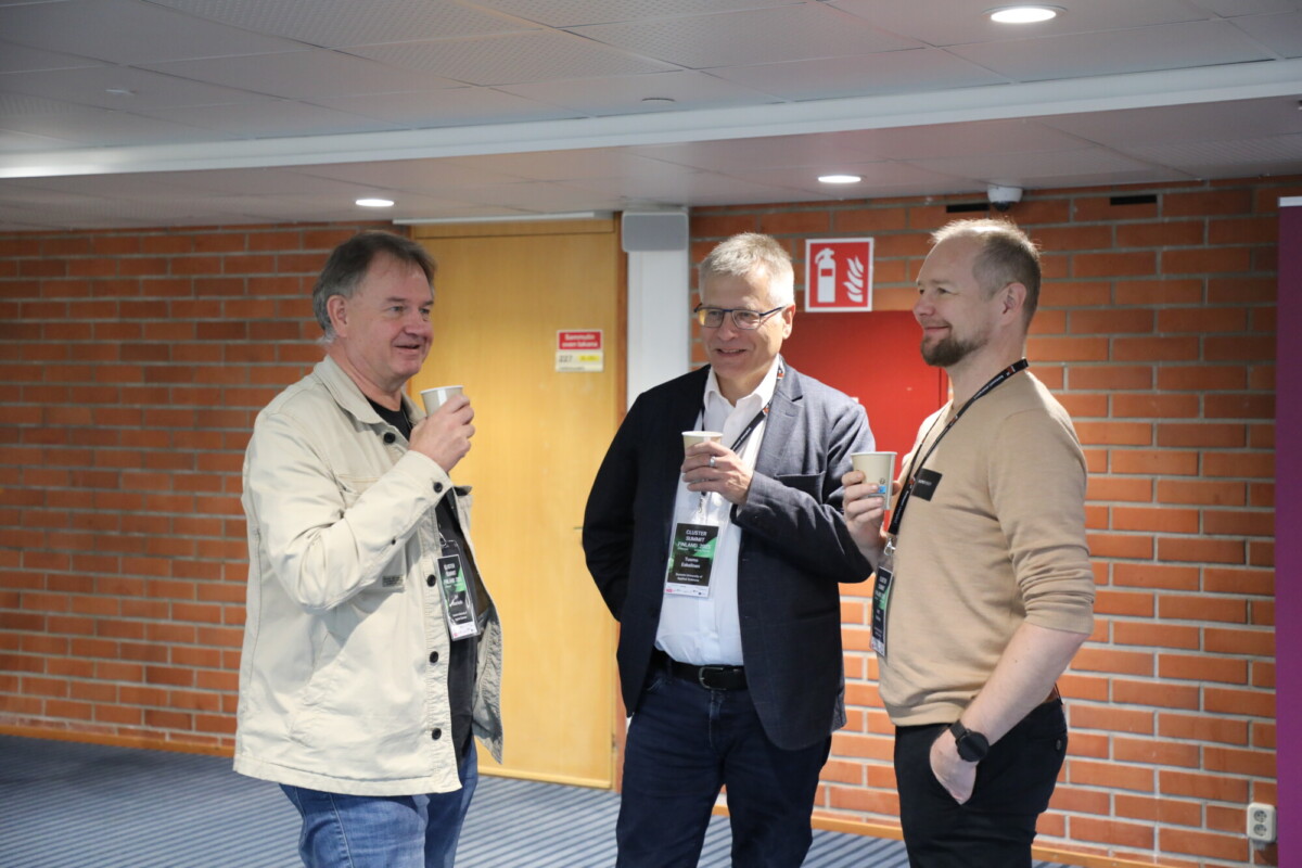 Kolme miestä seisoo käytävällä hymyillen ja kupit kädessä keskustellen. Kaikilla on konferenssimerkit. Taustalla on tiiliseinä, puinen ovi ja sammutinmerkki.