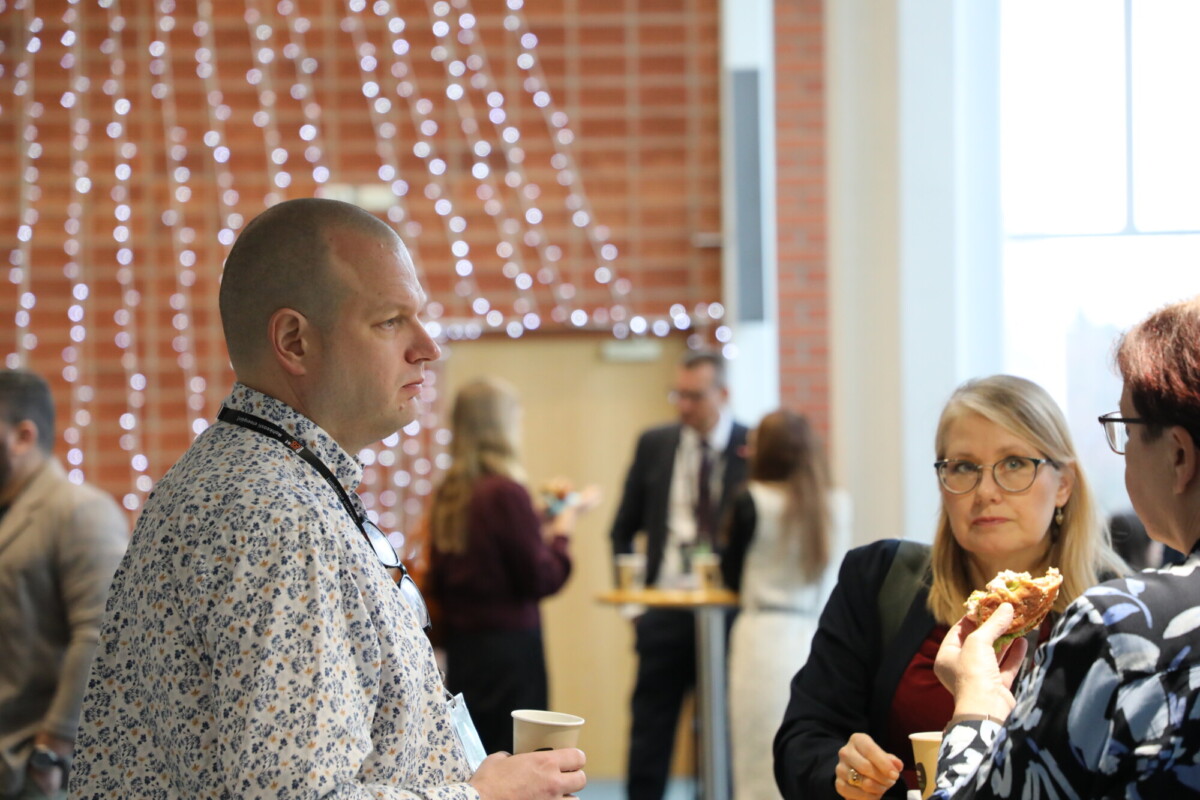 Kuppi kädessään pitävä mies keskustelee kahden naisen kanssa sisätiloissa järjestettävässä tapahtumassa. Taustalla on ihmisiä, ja tiiliseinää koristavat valonarut. Yksi nainen on syömässä, ja auringonvalo virtaa ikkunasta.