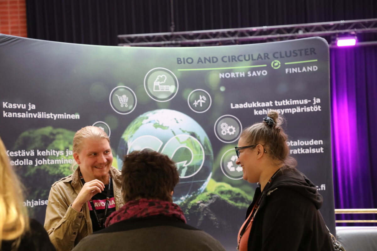 Kolme ihmistä puhuu Bio- ja ympyräklusteria esittelevän näytön edessä Pohjois-Savossa. Näytöllä on kuvia maapallosta, kuvakkeita ja tekstiä kestävästä kasvusta ja kehityksestä.
