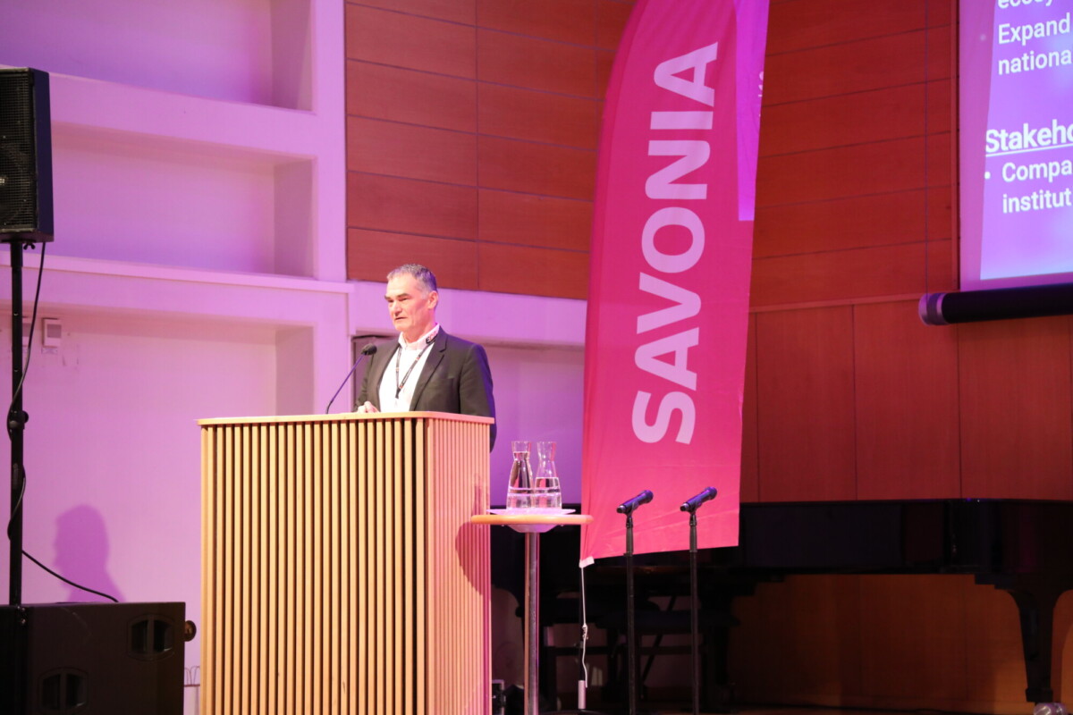 Pukumies seisoo puisella korokkeella puhumassa sisätiloissa järjestettävässä tilaisuudessa. Hänen takanaan on vaaleanpunainen banderolli, jossa lukee sana SAVONIA, ja suuri näyttö, jossa näkyy esityksen dia. Korokkeen edessä on kaksi mikrofonia.