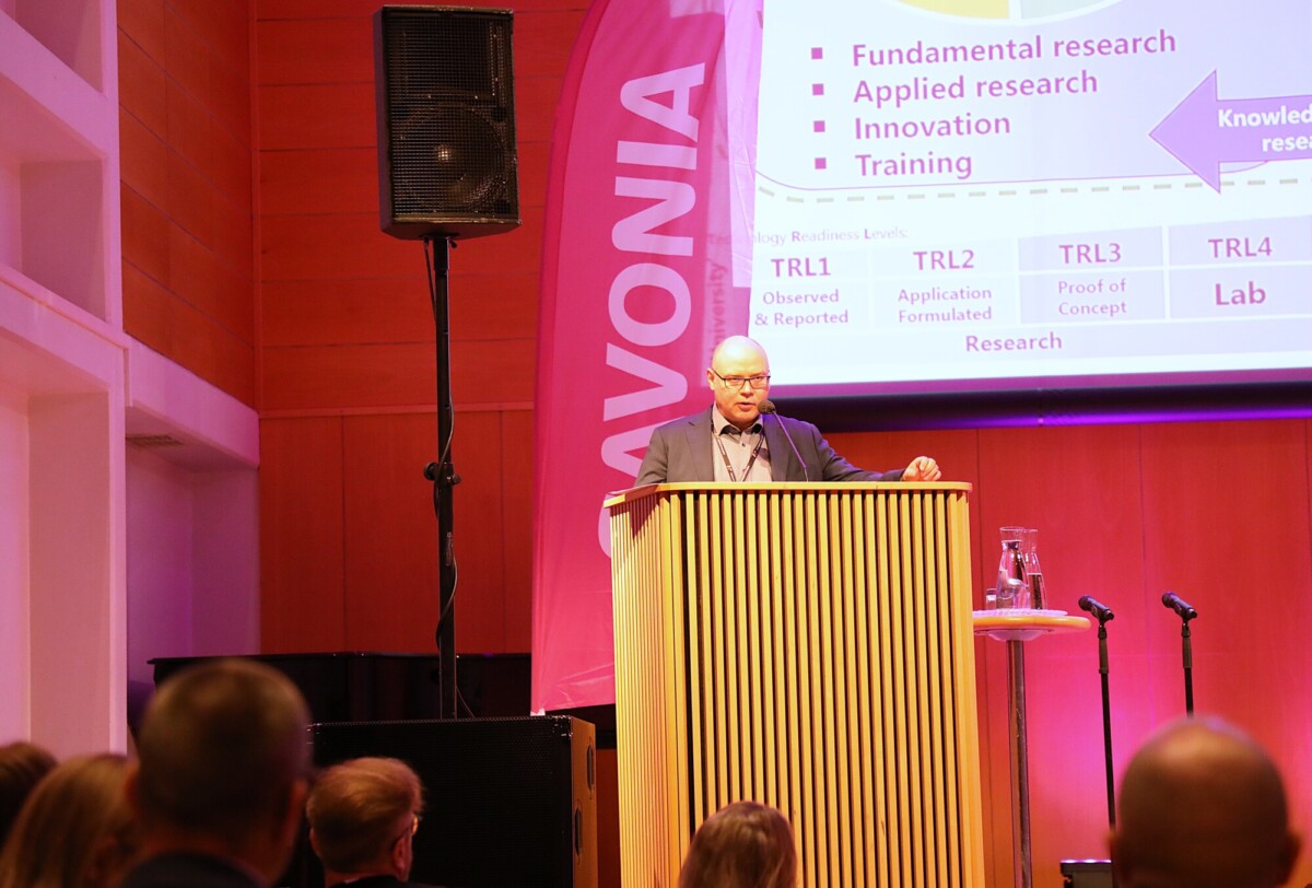 Mies seisoo korokkeella pitämässä esitelmää yleisölle. Hänen takanaan olevalla suurella näytöllä näkyy tekstiä tutkimuksesta ja innovaatiosta. Pystysuorassa bannerissa näkyy sana SAVONIA.