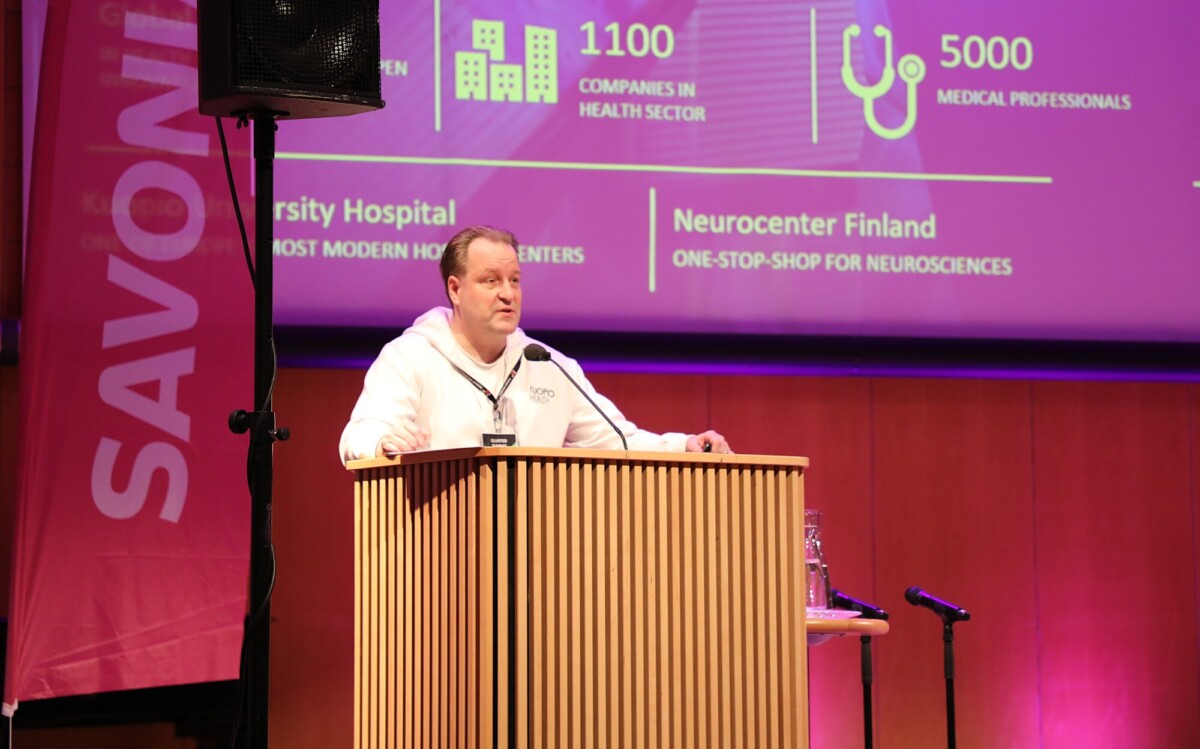 Valkoiseen huppariin pukeutunut mies puhuu puisella korokkeella lavalla. Hänen takanaan on suuri violetti esitysnäyttö, jossa on terveysalan tilastoja ja pystysuorassa bannerissa näkyvä Savonia-logo.