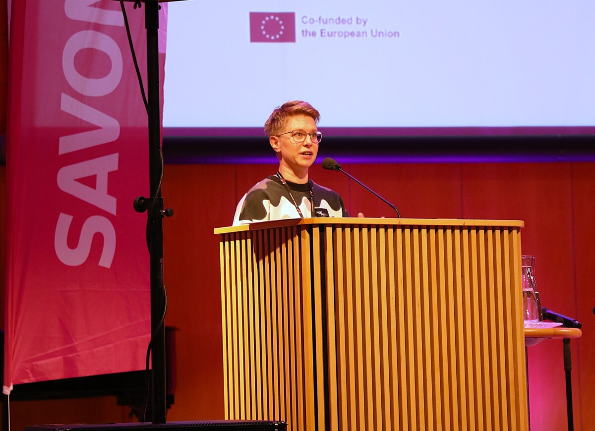 Lyhythiuksinen ja silmälasipäinen henkilö puhuu lavalla puisen korokkeen ääressä. Hänen takanaan on valkokangas, jossa on Euroopan unionin logo ja vaaleanpunainen banderolli, jossa näkyy osittain SAVON. Mikrofoni ja vesilasi ovat korokkeella.