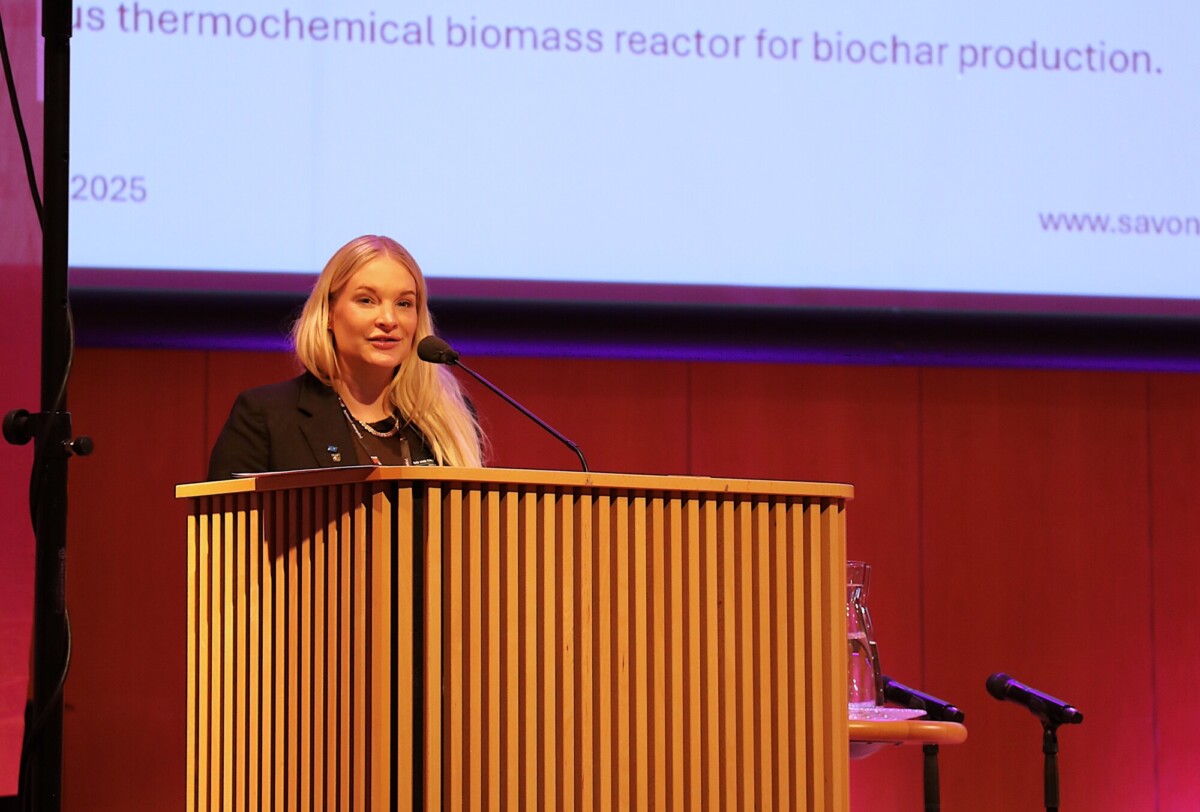 Pitkät vaaleat hiukset omaava nainen seisoo puisen korokkeen ääressä ja puhuu mikrofoniin, jonka edessä on heijastettu esitys biohiilen tuotantoon tarkoitetusta biomassareaktorista konferenssihuoneessa.