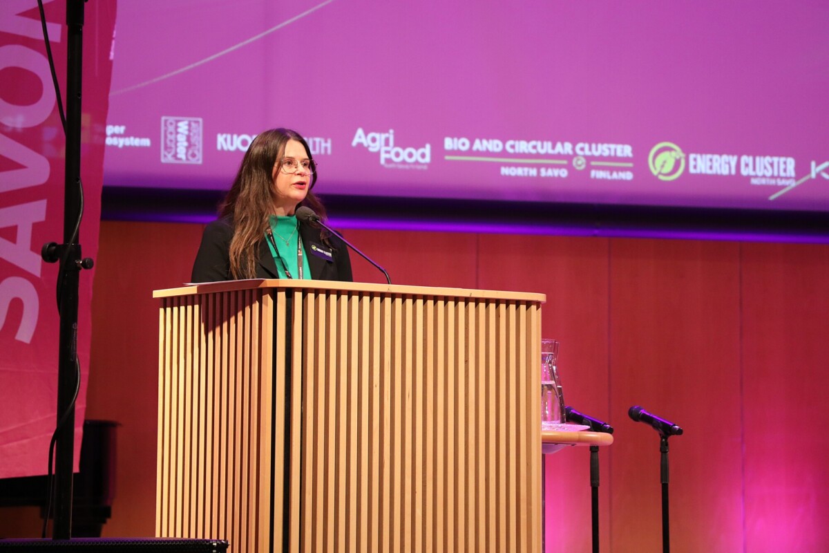 Pitkänruskeatukkainen nainen, jolla on silmälasit ja vihreä toppi mustan bleiserin alla, puhuu puisella korokkeella lavalla, jonka taustalla on violetti tausta, jossa näkyy tapahtuman logoja ja nimiä.