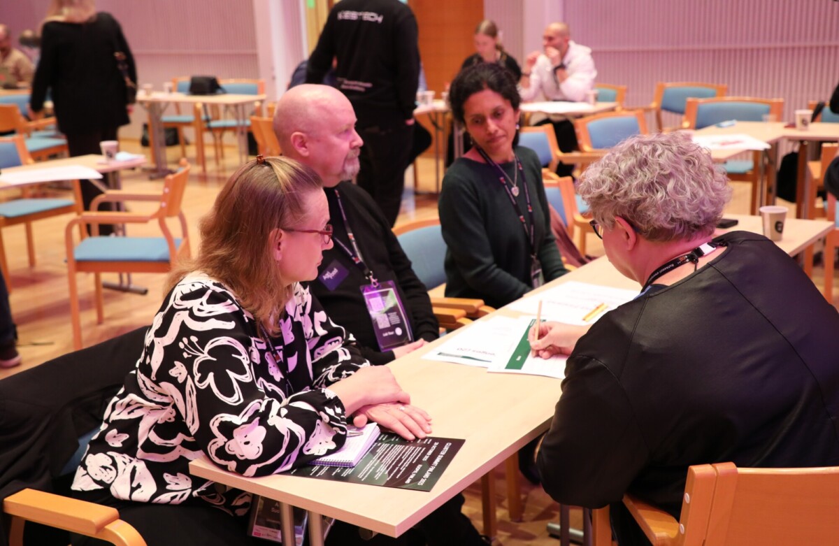 Neljä ihmistä istuu neuvotteluhuoneessa pöydän ympärillä keskustelemassa, paperit ja kynät edessään; taustalla näkyy muita osallistujia.