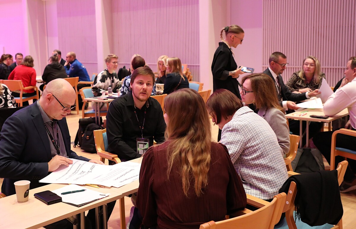 Ryhmä ihmisiä istuu pöytien ympärillä kirkkaasti valaistussa huoneessa keskustelemassa ja tekemässä muistiinpanoja, mikä viittaa työpajaan tai konferenssiin, jossa on yhteistoiminnallista toimintaa.