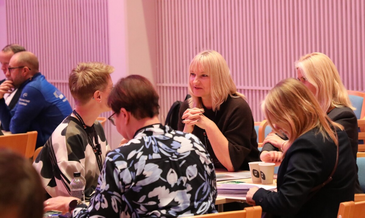 Joukko ihmisiä istuu pöydän ääressä kirkkaasti valaistussa huoneessa ja keskustelee. Pöydillä on papereita, muistikirjoja ja kuppeja, mikä viittaa kokous- tai työpajatilaisuuteen.