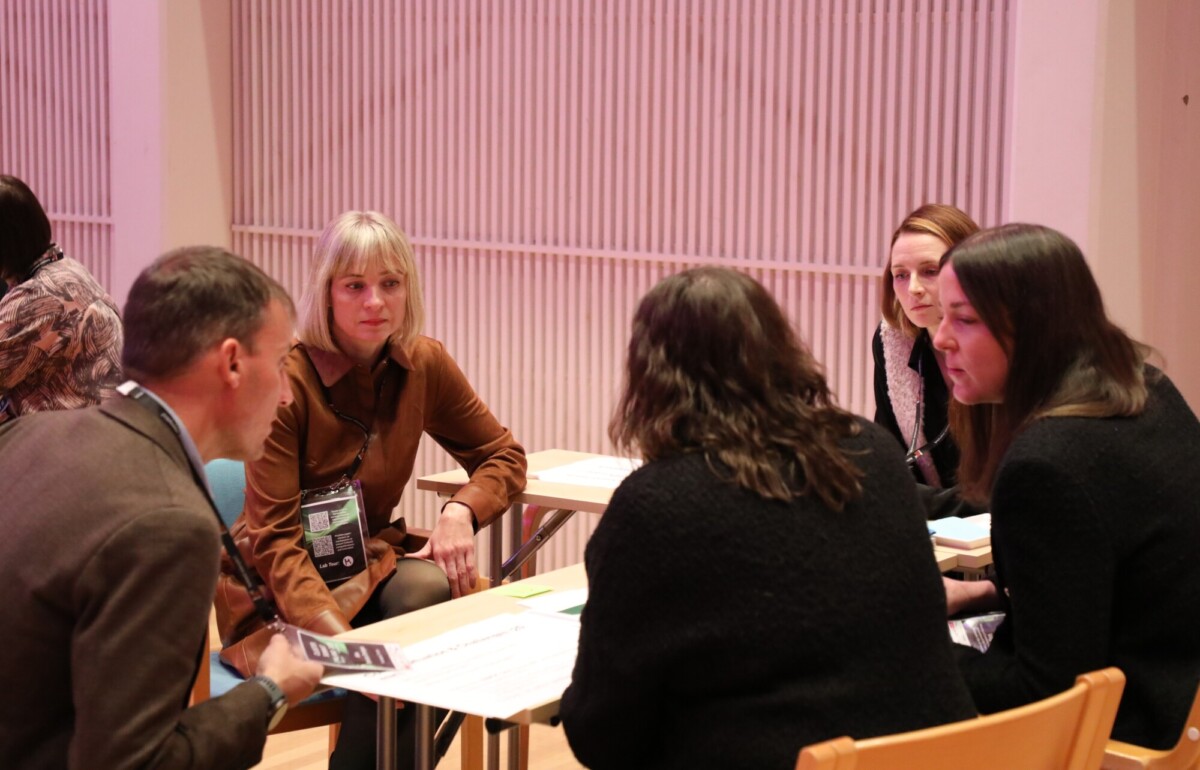 Viisi aikuista istuu piirissä sisätiloissa keskustelemassa. Heidän edessään olevalle pöydälle on levitetty papereita ja asiakirjoja. Ympäristö vaikuttaa muodolliselta, mahdollisesti kokoukselta tai työpajalta, ja taustalla on vaalea tausta.