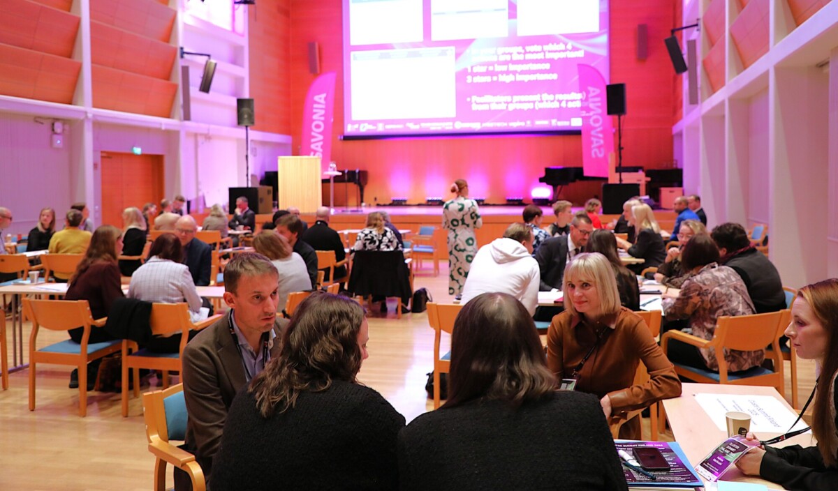 Joukko ihmisiä istuu pöydissä kirkkaasti valaistussa neuvotteluhuoneessa keskustelemassa. Suuri näyttö, jossa on vaaleanpunainen valaistus ja teksti, on huoneen etuosassa, mikä luo ammattimaisen ilmapiirin.