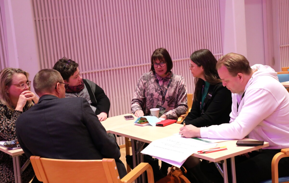 Kuusi aikuista istuu pöydän ääressä keskustelemassa, ja heidän edessään on papereita, muistilappuja ja värikyniä. He vaikuttavat keskittyneiltä ja tarkkaavaisilta hyvin valaistussa sisätilassa, jossa on vaaleat puutuolit.