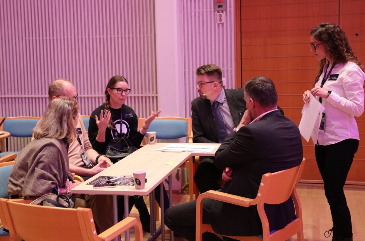 Kuusi ihmistä istuu ja seisoo pöydän ympärillä kirkkaasti valaistussa huoneessa keskustelemassa. Yksi nainen elehtii käsillään puhuessaan. Pöydällä on papereita, kuppeja ja kaulaliinoja; kaikki näyttävät olevan mukana.