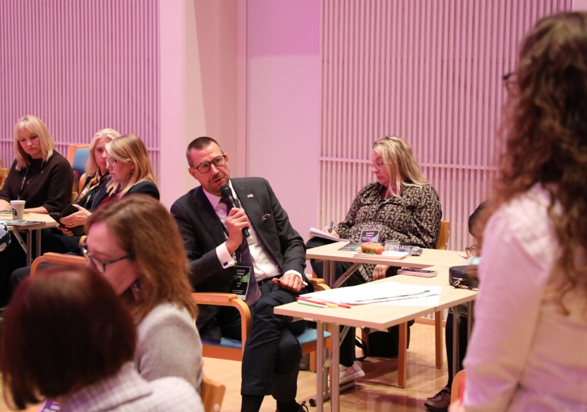 Pukumies puhuu mikrofoniin konferenssin tai seminaarin aikana, kun hän istuu muiden osallistujien joukossa pöytien ääressä ja nainen seisoo hänen edessään. Huoneessa on vaaleanpunainen valaistus ja pystysuorat valkoiset seinäpaneelit.