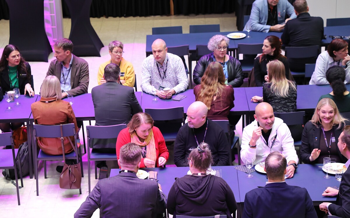 Joukko ihmisiä istuu tummansinisellä kankaalla peitettyjen pöytien ääressä keskustelemassa ja syömässä sisätiloissa järjestettävässä tapahtumassa tai konferenssissa. Useimmat keskustelevat, ja useilla on lautaset tai juomat edessään.