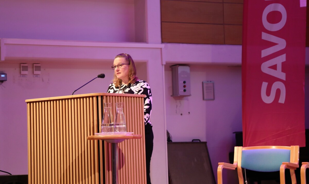 Nainen seisoo puisella korokkeella ja puhuu mikrofoniin kirkkaasti valaistussa huoneessa. Lähellä on suuri punainen banderolli, jossa on osittain teksti SAVO, ja tyhjä tuoli.