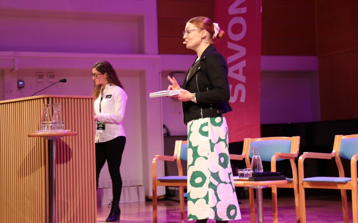 Kaksi naista seisoo lavalla; toinen puhuu kuulokemikrofoniin ja pitää kädessään muistikirjaa, kun taas toinen seisoo korokkeen vieressä. Taustalla on neljä tyhjää tuolia ja suuri vaaleanpunainen banderolli, jossa lukee SAVON.