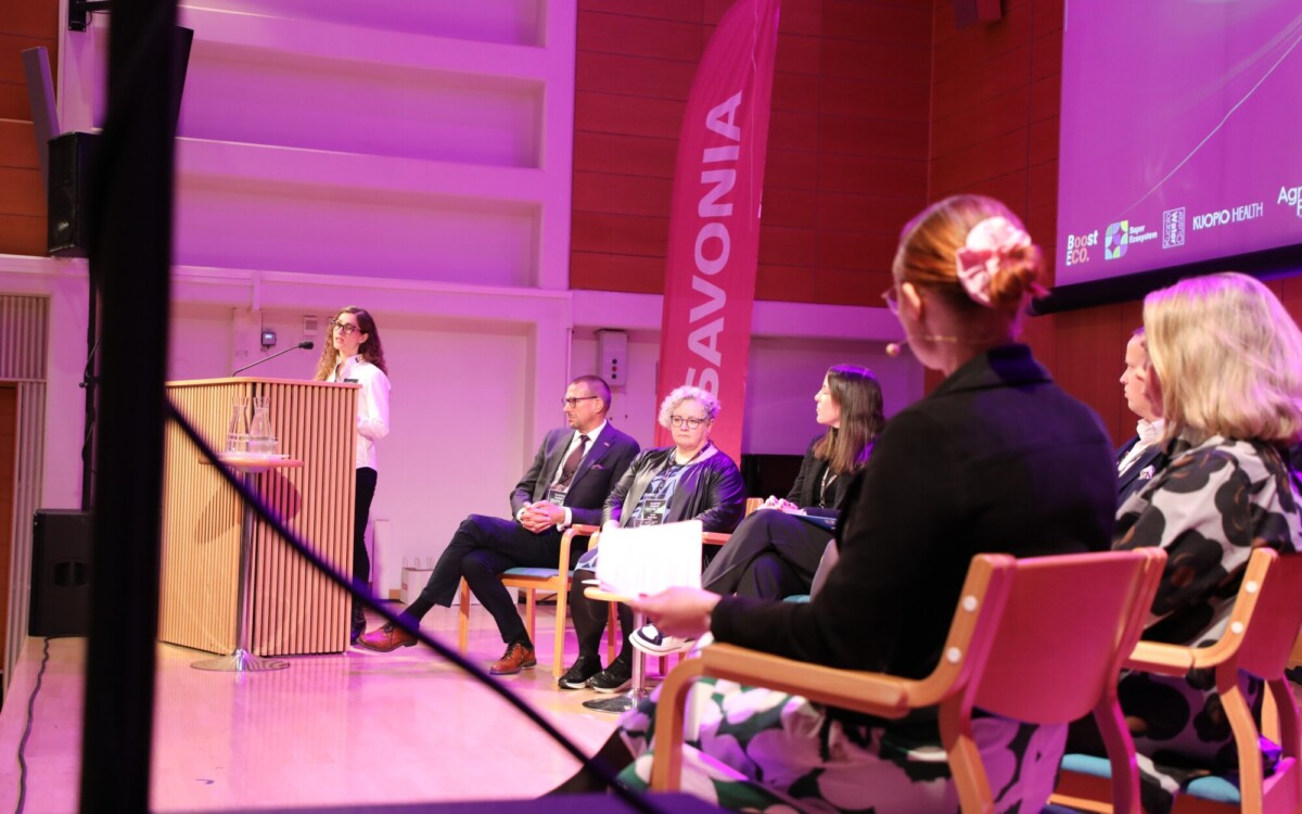 Nainen puhuu lavalla korokkeella viiden istuvan panelistin kuunnellessa. Tilanne on sisätiloissa, joissa on violetti valaistus, ja taustalla näkyy pystysuora banderolli, jossa lukee SAVONIA.