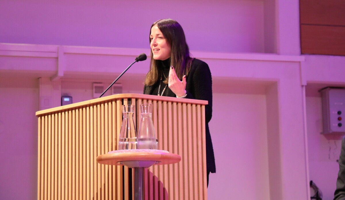 Pitkät tummat hiukset omaava, mustiin pukeutunut nainen puhuu puisella korokkeella, jossa on mikrofoni. Korokkeen edessä olevalla pienellä pyöreällä pöydällä on kaksi lasista vesikannua ja lasi. Huone on valaistu violetilla valolla.