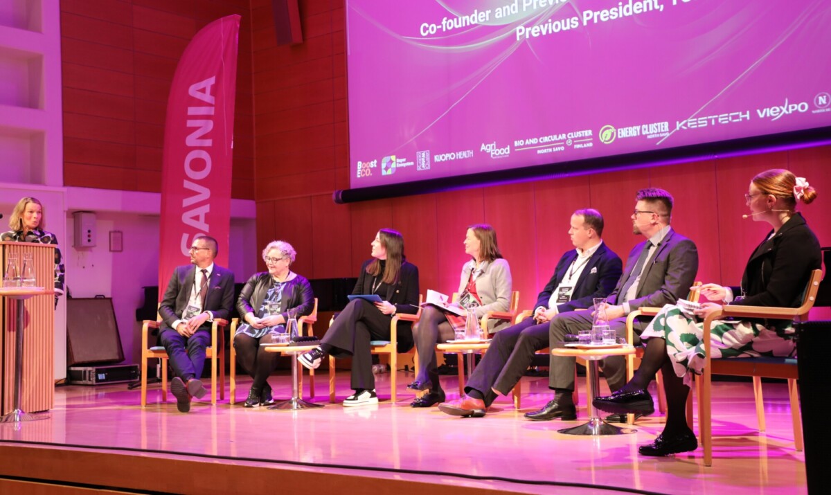 Seitsemän hengen paneeli istuu lavalla tuoleilla yleisöön päin. Nainen seisoo korokkeella puhumassa. Heidän takanaan on suuri violetti näyttö, jossa näkyy tapahtumaa ja sponsoreita koskevia tietoja.