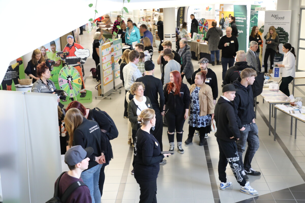 Vilkas sisätiloissa järjestetty tapahtuma, jossa ihmiset kerääntyivät osastojen ja näytteiden ympärille. Jotkut keskustelevat, toiset katselevat näyttelyesineitä. Ilmapiiri on vilkas ja sosiaalinen, ja taustalla näkyy tiedotusmateriaalia ja julisteita.