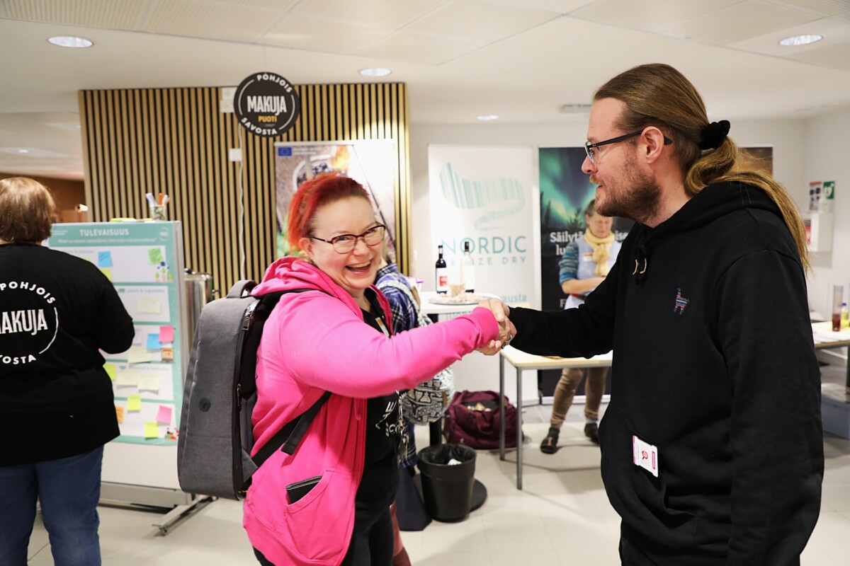 Kaksi ihmistä hymyilee ja kättelee toisiaan sisätiloissa, joiden takana on tiedotuskoppeja ja julisteita. Toisella on vaaleanpunainen huppari ja reppu, toisella silmälasit ja musta huppari.