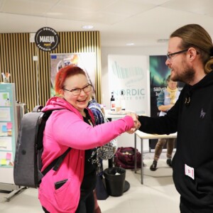 Kaksi ihmistä hymyilee ja kättelee toisiaan sisätiloissa, joiden takana on tiedotuskoppeja ja julisteita. Toisella on vaaleanpunainen huppari ja reppu, toisella silmälasit ja musta huppari.