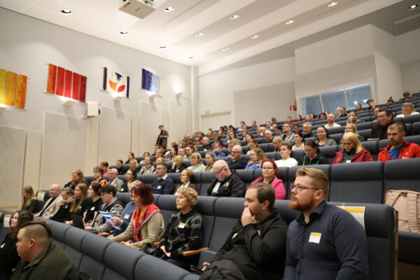 Suuri joukko ihmisiä istuu luentosalissa istuinrivissä ja katselee tarkkaavaisesti jotakin edessä olevaa asiaa. Yleisö on iältään ja sukupuoleltaan moninainen, ja seinällä roikkuu värikkäitä julisteita.