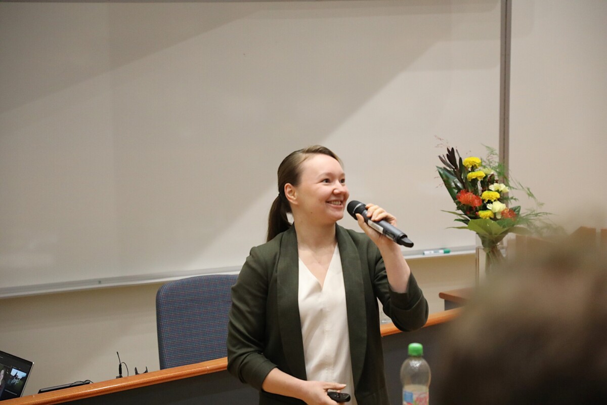 Vihreään bleiseriin pukeutunut nainen seisoo luentokorokkeella hymyillen ja mikrofoni kädessään. Hänen takanaan on valkotaulu, kukkakimppu ja vesipullo pöydällä.