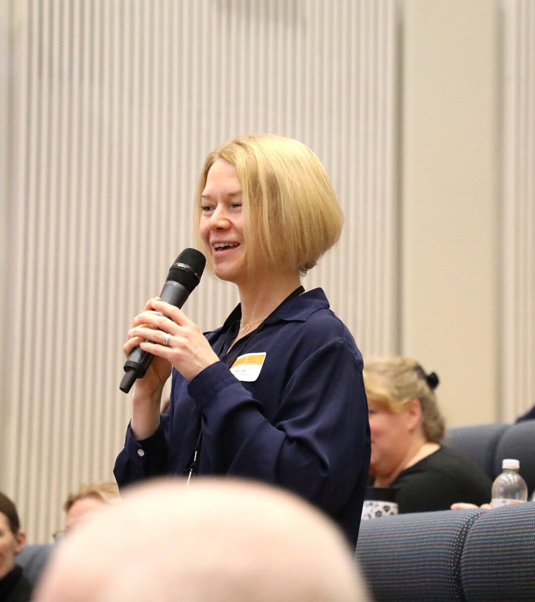 Lyhyet vaaleat hiukset omaava nainen seisoo auditoriossa mikrofoni kädessään ja puhuu, kun hänen ympärillään istuvat ihmiset kuuntelevat tarkkaavaisesti.