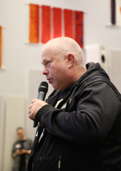 Lyhyet, vaaleat hiukset ja musta takki päällään oleva henkilö puhuu mikrofoniin sisätiloissa. Taustalla seinällä roikkuu värikkäitä kangasbanderolleja.