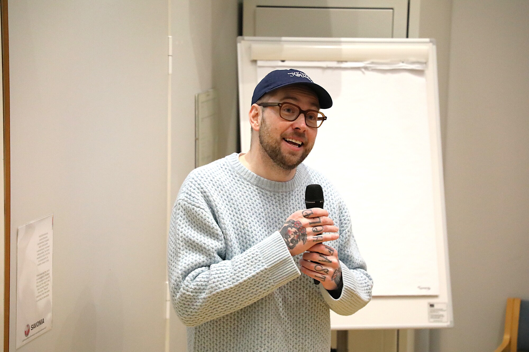 Henkilö, jolla on silmälasit, sininen lippis ja vaaleansininen villapaita, pitää mikrofonia kädessään puhuessaan. He seisovat sisätiloissa tyhjän fläppitaulun vieressä. Heidän käsissään näkyy tatuointeja.