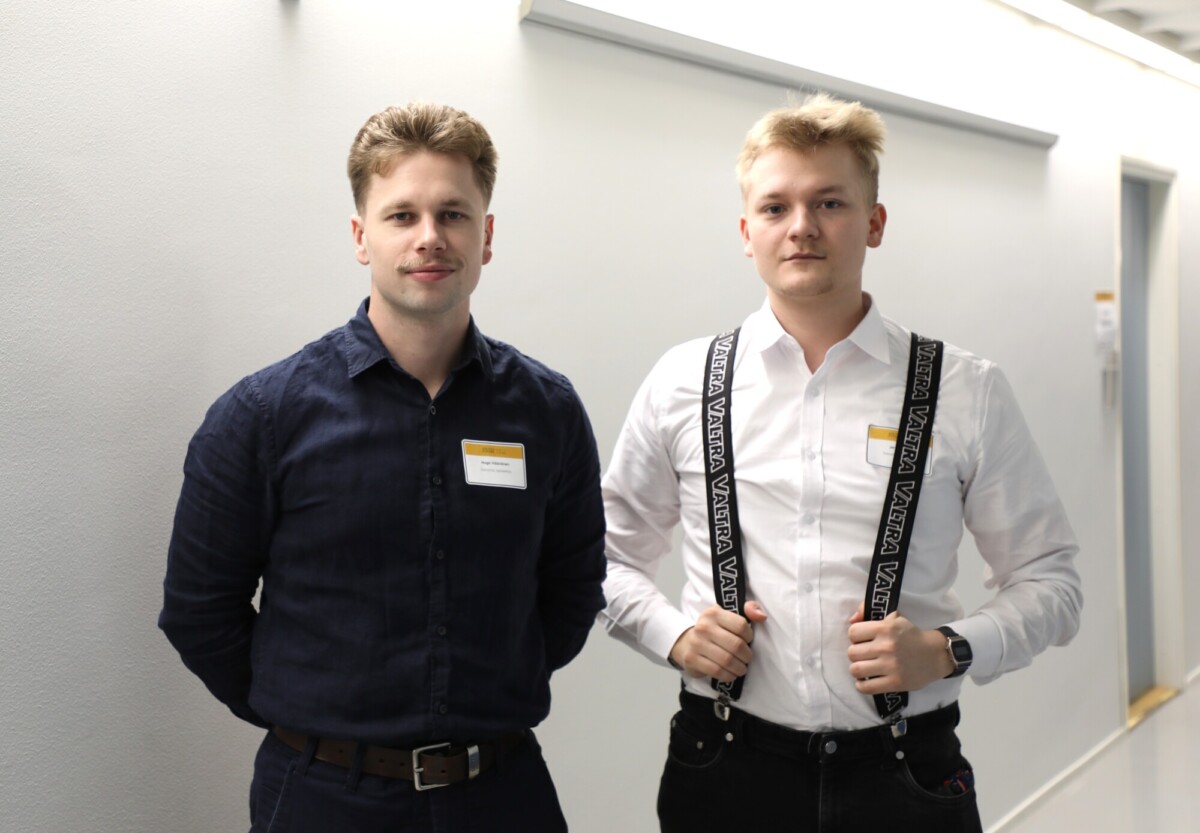 Kaksi nuorta miestä seisoo sisätiloissa valkoista seinää vasten. Toisella on tumma paita ja ruskea vyö, toisella valkoinen paita ja mustat housunkannattimet, joissa on merkintä Vans. Molemmilla on vaaleat hiukset, ja heillä on nimilaput rinnassaan.