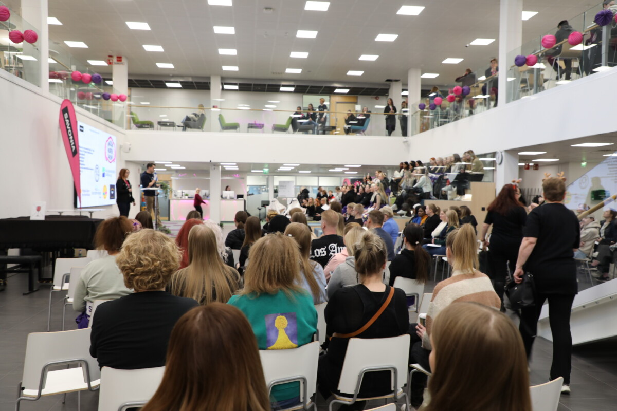 Suuri joukko ihmisiä istuu modernissa, valoisassa kaksikerroksisessa salissa kuunnellen puhujia lavalla. Kaiteita koristavat vaaleanpunaiset ilmapallot, ja osa osallistujista seisoo tai istuu ylemmällä tasolla.