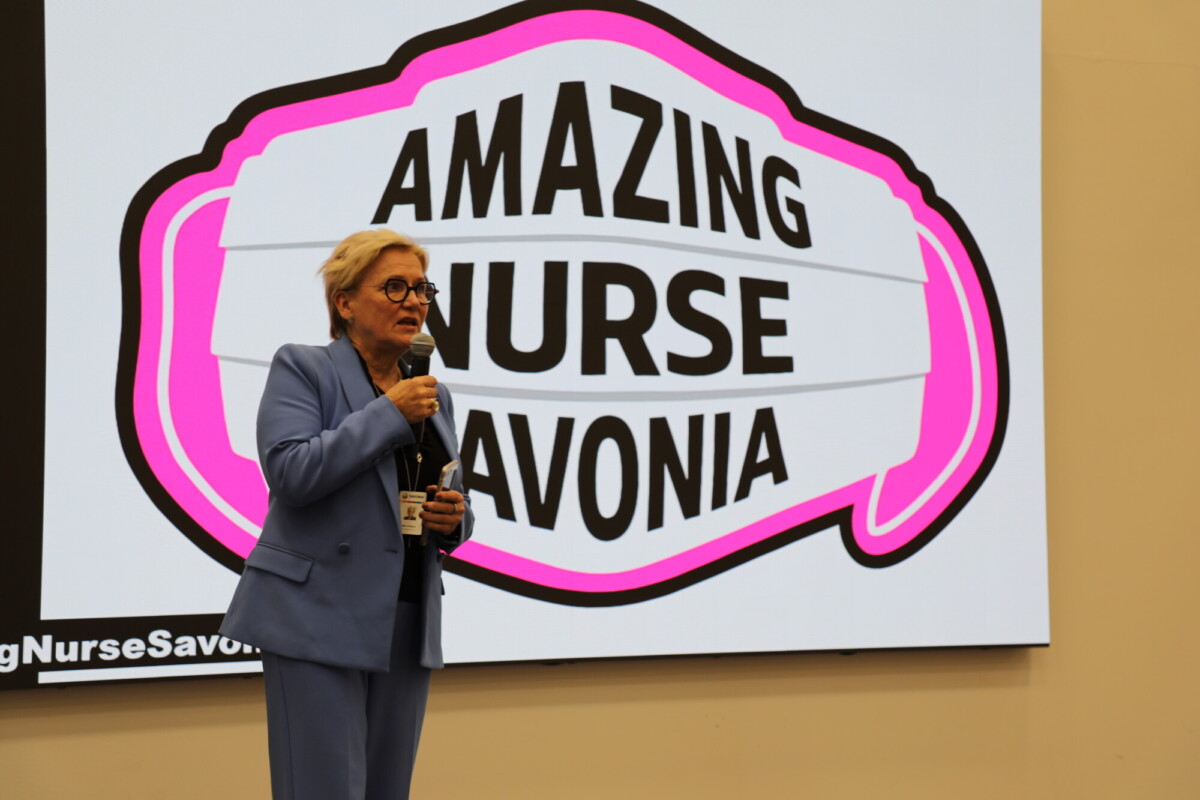 Vaaleansiniseen pukuun pukeutunut henkilö puhuu mikrofoniin näytön edessä, jossa on vaaleanpunaisen ja valkoisen puhekuplan sisällä teksti AMAZING NURSE SAVONIA.