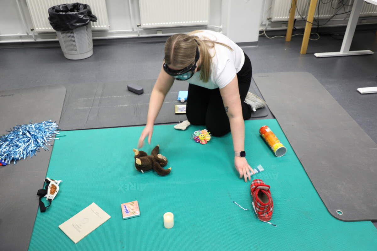 Henkilö, jolla on silmäside, polvistuu vihreällä jumppamatolla ja kurottautuu kohti ruskeaa pehmolelua. Erilaisia esineitä, kuten kynttilä, kirja ja hyppynaru, on ripoteltu matolle heidän ympärilleen.