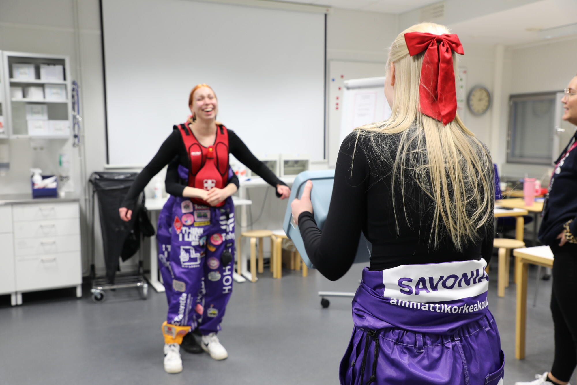 Kaksi nuorta naista, joilla on purppuranpunaiset haalarit, jotka on peitetty laastareilla ja asusteilla, seisovat luokkahuoneessa hymyillen ja vuorovaikutuksessa. Toisella on suuri punainen rusetti hiuksissaan, ja huoneessa on hoitotarvikkeita ja puutuoleja.