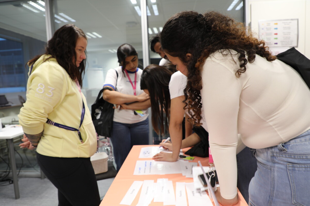 Useita nuoria naisia seisoo nimilappujen ja papereiden peittämän pöydän ympärillä kirkkaasti valaistussa huoneessa. Yksi nainen kirjoittaa, kun muut katselevat ja odottavat, mikä viittaa rekisteröinti- tai ilmoittautumisprosessiin.