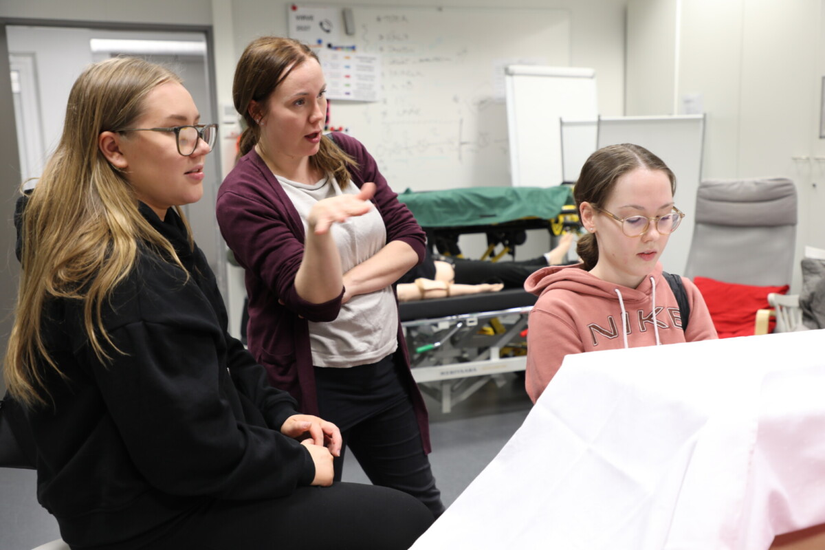 Kolme naista on lääketieteellisessä koulutushuoneessa. Yksi puhuu eleillä, kun kaksi muuta kuuntelevat. Taustalla näkyy lääketieteellisiä laitteita ja mallinukkeja, ja seinällä on valkotaulu.