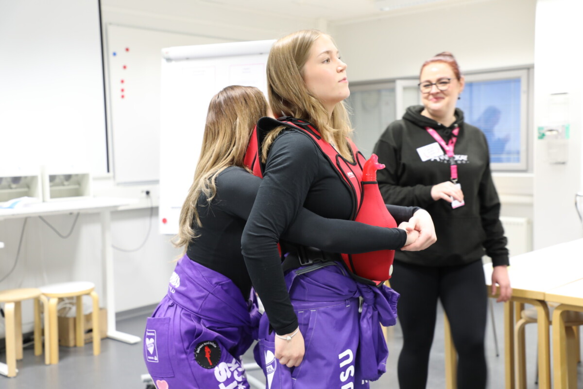 Kaksi nuorta naista, joilla on samanlaiset violetit haalarit ja mustat paidat, harjoittelevat pelastusnostotekniikkaa punaisilla valjailla, kun taas toinen mustapukuinen nainen seisoo lähellä katsomassa ja hymyilemässä kirkkaassa luokkahuoneessa.