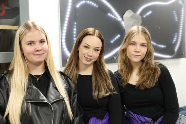 Kolme pitkätukkaista nuorta naista poseeraa sisätiloissa, kaikilla yllään mustat topit. Kahdella oikealla olevalla on myös violetit liivit. Heidän takanaan olevalla seinällä näkyy moderni taideteos, jossa on valoja.